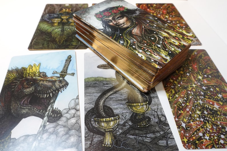 May include: A set of tarot cards with colorful illustrations. The cards feature a variety of images, including a dinosaur, a woman with flowers in her hair, and a snake with a cup. The cards are arranged in a fan shape on a white surface.