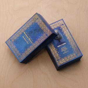May include: Two blue boxes with gold trim and intricate designs. The top box has the text "Oceanum Arcanae The Ocean Tarot" printed on it. The bottom box has the text "The Sun" printed on it.