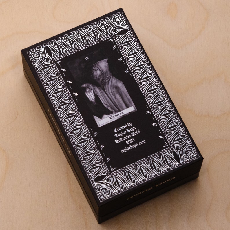 May include: Black box with a silver border and a tarot card image of The Hermit. The card is black and white and features a hooded figure holding a lantern. The text on the card reads "IX The Hermit" and "Created by Taylor Bryn Illustrator-Todd 2021 taylorbryn.com".