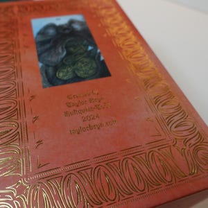 May include: A red box with gold embossed details and a small image of a creature on the front. The box has the text "Taylor Bey" and "Bughouse-Tees 2023" on the front.
