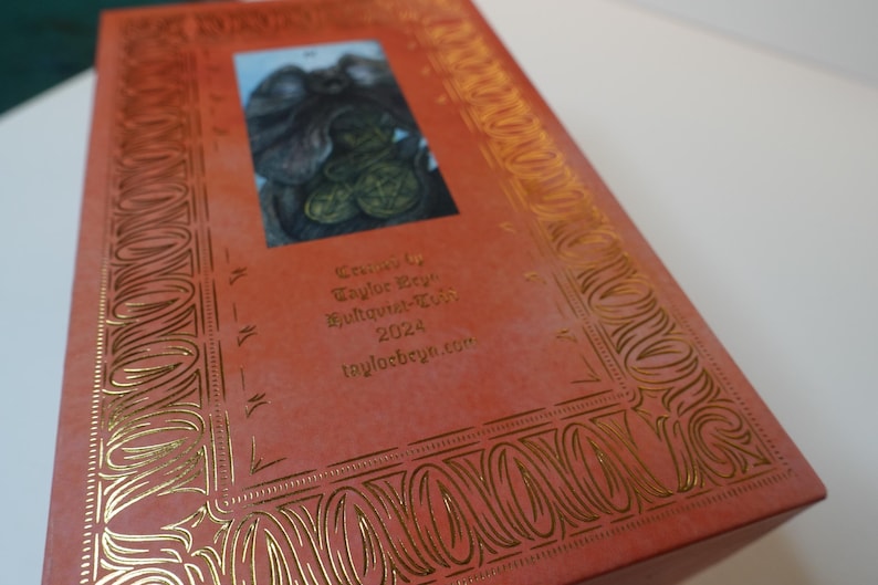 May include: A red box with gold embossed details and a small image of a creature on the front. The box has the text "Taylor Bey" and "Bughouse-Tees 2023" on the front.