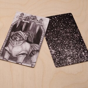 May include: A black and white tarot card with a dog wearing armor and a sword in its mouth. The card is labeled "IV" and has a black and white background with a pattern of stars.