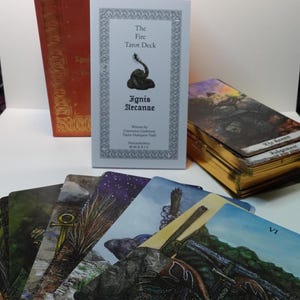 May include: A tarot deck box with a gold trim and a snake design on the cover. The box is closed and the title "The Fire Tarot Deck" is visible on the cover. The text "Ignis Arcanae" is also visible on the cover. The box is sitting on a table with a stack of tarot cards in the background.