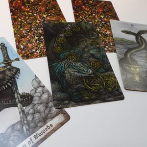 May include: Four tarot cards, each with a different illustration. The first card shows a green and brown lizard with a sword through its head. The second card shows a green and brown lizard with gold coins. The third card shows a green and brown lizard with gold coins. The fourth card shows two snakes intertwined with gold coins.