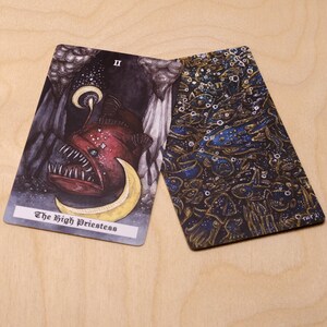 May include: Two tarot cards, one with a black background and a red and white anglerfish with a crescent moon, and the other with a blue and gold background with a pattern of fish and bubbles. The card with the anglerfish has the text "The High Priestess" and the Roman numeral "II" on it.
