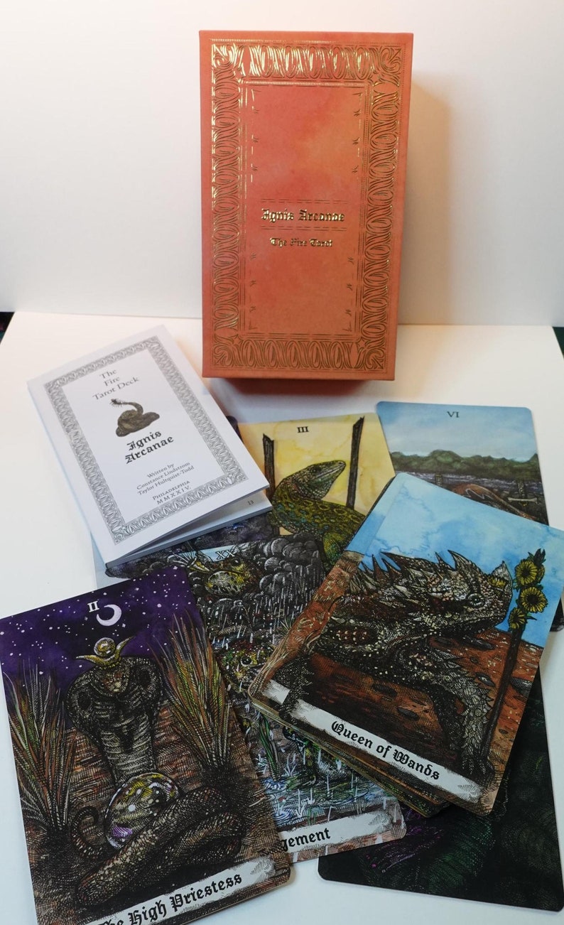 May include: A tarot deck box with a red cover and gold lettering that reads "Agnis Arcanae The Fire Tarot". The box is open and shows a white booklet with the title "The Fire Tarot Deck Ignis Arcanae" and several tarot cards with illustrations of lizards and other creatures.