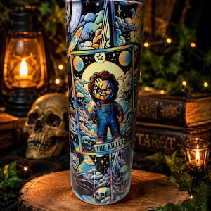 May include: A cylindrical tumbler featuring a colourful graphic print of a doll in blue overalls holding a knife, framed within a tarot card design labelled THE KILLER. The artwork includes stylised clouds, stars, and skulls in vibrant hues.