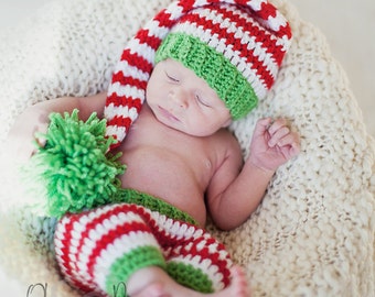 May include: A newborn baby wearing a red, white and green striped crocheted elf hat and matching trousers. The baby is sleeping peacefully in a white crocheted basket.