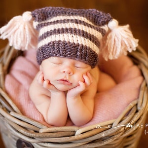 Puede incluir: Un bebé recién nacido con un gorro de punto a rayas morado y blanco con pompones rosas, durmiendo en una cesta de mimbre forrada con una manta rosa.