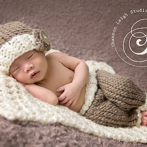 May include: A newborn baby wearing a brown and white knitted hat with a button and brown knitted pants is sleeping on a white and brown knitted blanket.