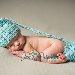 May include: A newborn baby wearing a blue and white crocheted hat with a long tail and a matching pom-pom. The baby is sleeping on a light grey surface.