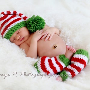 May include: A newborn baby wearing a red and green striped knit hat with a green pom-pom and matching red and green striped leg warmers.