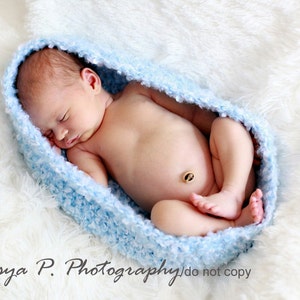 May include: A newborn baby sleeping in a light blue crocheted baby nest on a white fluffy surface.