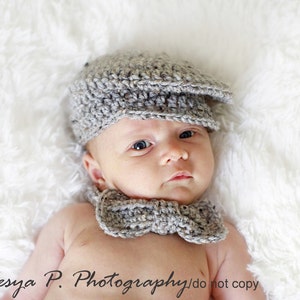 May include: A newborn baby wearing a gray crocheted hat and bow tie. The baby is lying on a white fluffy surface.