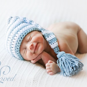 May include: A newborn baby wearing a blue and white crocheted hat with a long tassel. The baby is lying on a white blanket and has their eyes closed.