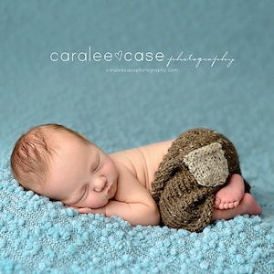 Puede incluir: Un bebé recién nacido está durmiendo en una manta azul texturizada. El bebé lleva un pañal de punto marrón con un parche beige en la espalda.