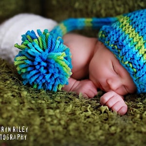 May include: A sleeping baby wearing a blue and green knitted hat with a blue pom-pom. The baby is lying on a green fuzzy surface.