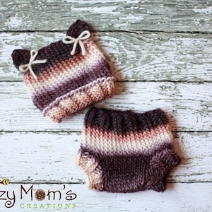 May include: A knitted hat and diaper set in a brown, pink, and white striped pattern. The hat has cat ears and bows. The set is on a white wooden background.