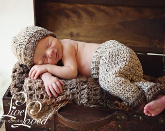 May include: A newborn baby wearing a crocheted hat and trousers sleeps in a wooden trunk. The baby is lying on a brown fluffy blanket. The trunk is decorated with a brown leather handle and metal accents.