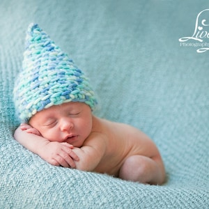 May include: A newborn baby wearing a blue and white knitted hat sleeps on a light blue blanket. The baby is curled up with their arms around their face.
