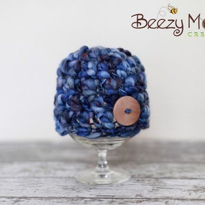 Puede incluir: Un gorro de bebé de crochet azul y morado con un botón marrón. El gorro se muestra en un pedestal de vidrio transparente.