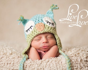 May include: A newborn baby wearing a green and blue crocheted owl hat with black eyes and a brown nose. The baby is sleeping with their hands under their chin. The baby is lying on a white fluffy blanket.