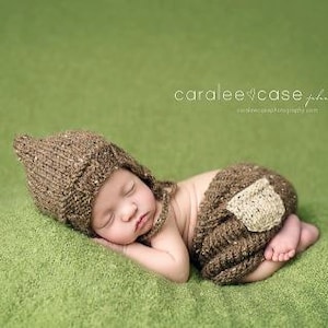 Puede incluir: Un bebé recién nacido con un gorro de punto marrón y pantalones a juego con un parche marrón y beige en la pierna, durmiendo sobre una superficie verde.