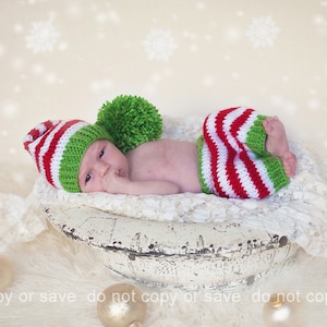 Puede incluir: Un bebé recién nacido con un gorro de punto a rayas verdes y rojas con un pompón verde y pantalones de punto a rayas verdes y rojas a juego. El bebé está acostado en un cuenco blanco sobre una superficie blanca y peluda.