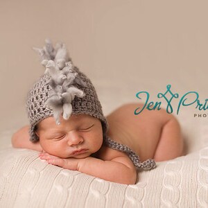 May include: A newborn baby wearing a gray crocheted hat with a gray and white fuzzy pom-pom on top, sleeping on a white knitted blanket. The baby is wearing a gray and white crocheted hat with a gray and white fuzzy pom-pom on top.