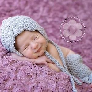 Puede incluir: Un bebé con un gorro y botines de ganchillo grises. El bebé está acostado sobre una manta suave, rosa y esponjosa.