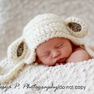 May include: A newborn baby wearing a white crocheted lamb hat with brown felt ears. The baby is sleeping on a white textured blanket.