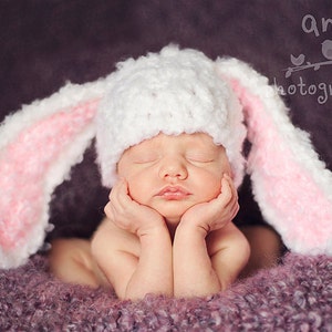 May include: A baby wearing a fluffy white bunny hat with pink inner ears, lying on a purple blanket. The baby has their eyes closed and hands under their chin. The image has a soft focus, with the text "am photography" in the top right corner.