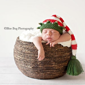 May include: A newborn baby wearing a green and red striped elf hat with jingle bells and a green pom-pom sleeps in a brown wicker basket.