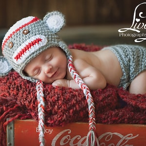 May include: A sleeping baby wearing a gray and white crocheted monkey hat and matching diaper cover. The baby is lying on a red fuzzy blanket in a wooden crate with the word "Enjoy" printed on the side.
