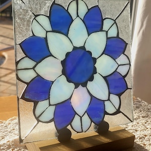 May include: A stained glass window panel featuring a blue and white flower design. The panel is mounted on a wooden stand.