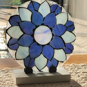 May include: A stained glass flower with blue and white petals on a white wooden base. The flower is round and has a white center.