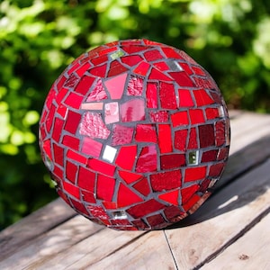 May include: A decorative red mosaic glass gazing ball. The sphere is composed of various shades of red glass tiles, creating a textured surface. The mosaic pieces are arranged in a geometric pattern, with dark gray grout lines. The ball is sitting on a wooden surface.