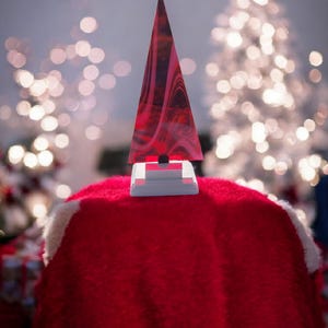 May include: A decorative Christmas tree with a red and black swirled design. The tree is set on a white base and sits on a red, fuzzy surface. The background features blurred Christmas lights.