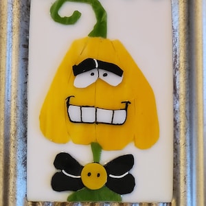 May include: A whimsical, rectangular glass art piece featuring a smiling yellow flower with a green stem and a black bow tie. The flower has white eyes and a wide, toothy grin. The background is white, and the frame is silver.