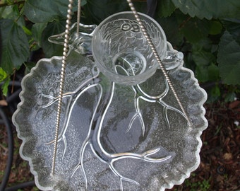 May include: A clear glass bird feeder with a decorative leaf-shaped base and a small cup for water or seeds. The feeder hangs from a silver chain and is surrounded by green foliage.
