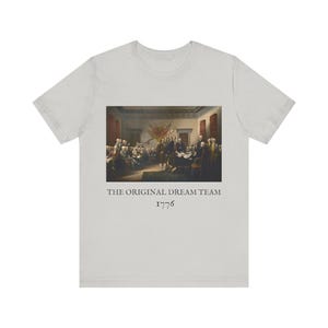 May include: A light gray t-shirt featuring a historical painting of a group of men. The text "THE ORIGINAL DREAM TEAM 1776" is printed below the image in a simple font. The shirt has short sleeves and a crew neck.