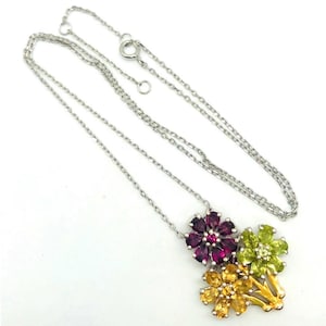 May include: A silver-toned chain necklace featuring a pendant with three flower-shaped clusters. The flowers are set with faceted gemstones in deep magenta, light green, and golden yellow, with a small central stone in each flower.