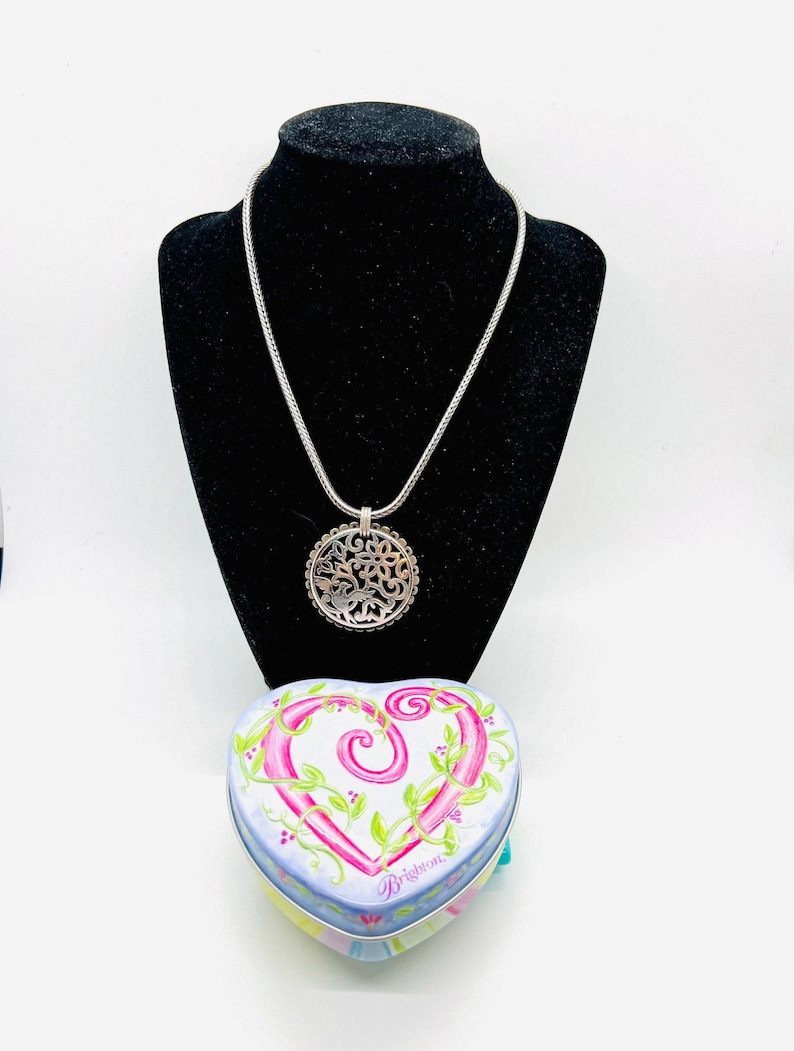 May include: A silver necklace with a delicate chain and a circular pendant featuring a floral design. The necklace is displayed on a black velvet stand. A heart-shaped tin box with a floral design and the word "Brighton" is also shown.
