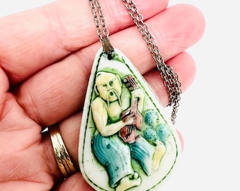 May include: A vintage pendant necklace with a teardrop-shaped pendant. The pendant features a carved figure playing a stringed instrument, with green, blue, and yellow accents. The necklace has a silver-toned chain.