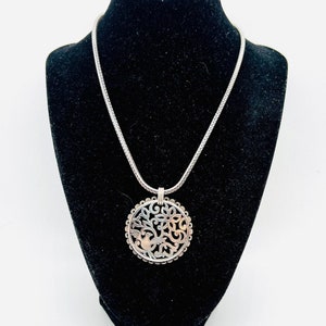 May include: A silver necklace with a round pendant featuring a delicate, cut-out design of a bird in a tree. The pendant has a two-tone finish with silver and gold accents.