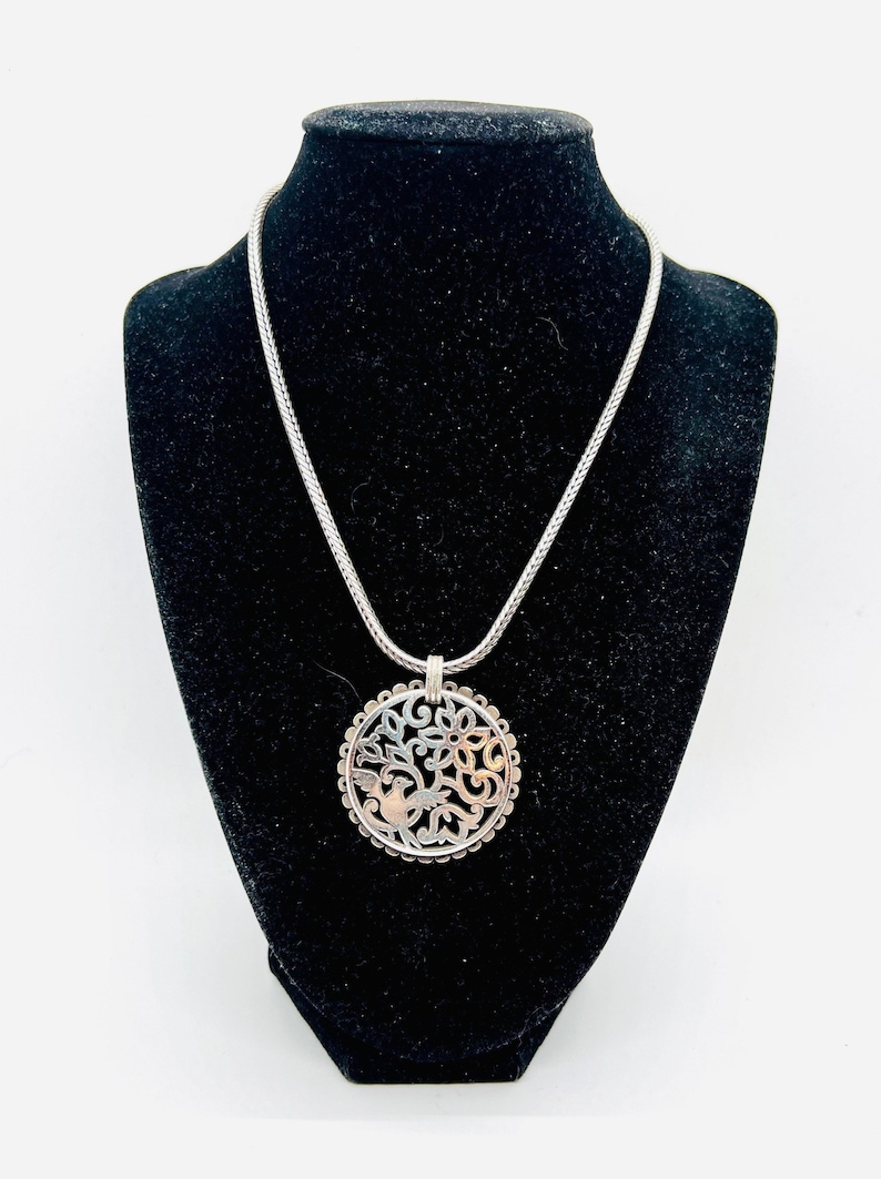 May include: A silver necklace with a round pendant featuring a delicate, cut-out design of a bird in a tree. The pendant has a two-tone finish with silver and gold accents.