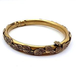 May include: A gold-toned hinged bangle bracelet. The bracelet is adorned with a repeating leaf and vine pattern. A small flower clasp secures the bracelet. The jewelry is set against a white background.