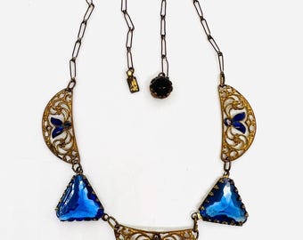 May include: A gold necklace with a geometric design featuring blue and black glass stones. The necklace has a chain with a clasp and a small black stone pendant.