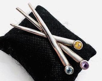 May include: A silver brooch with three prongs, each topped with a gemstone. The gemstones are blue, yellow, and purple.