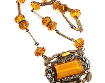 May include: Vintage amber-colored necklace with a rectangular pendant. The pendant is framed by an ornate, bronze-toned metal design with floral and leaf accents. The necklace features amber-colored faceted beads and bronze-toned metal links.
